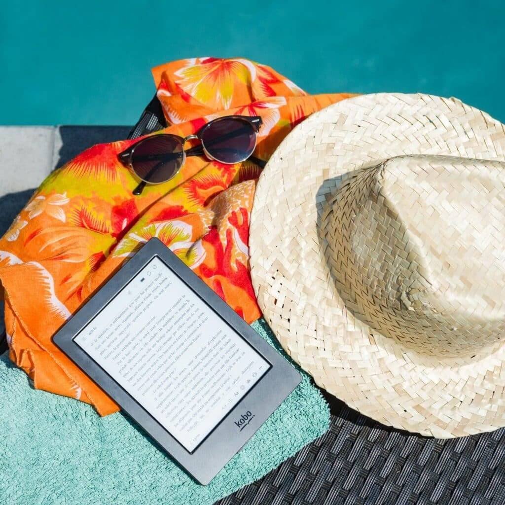 Flat lay of summer essentials by the pool with e-reader, sunhat, and vibrant towel.