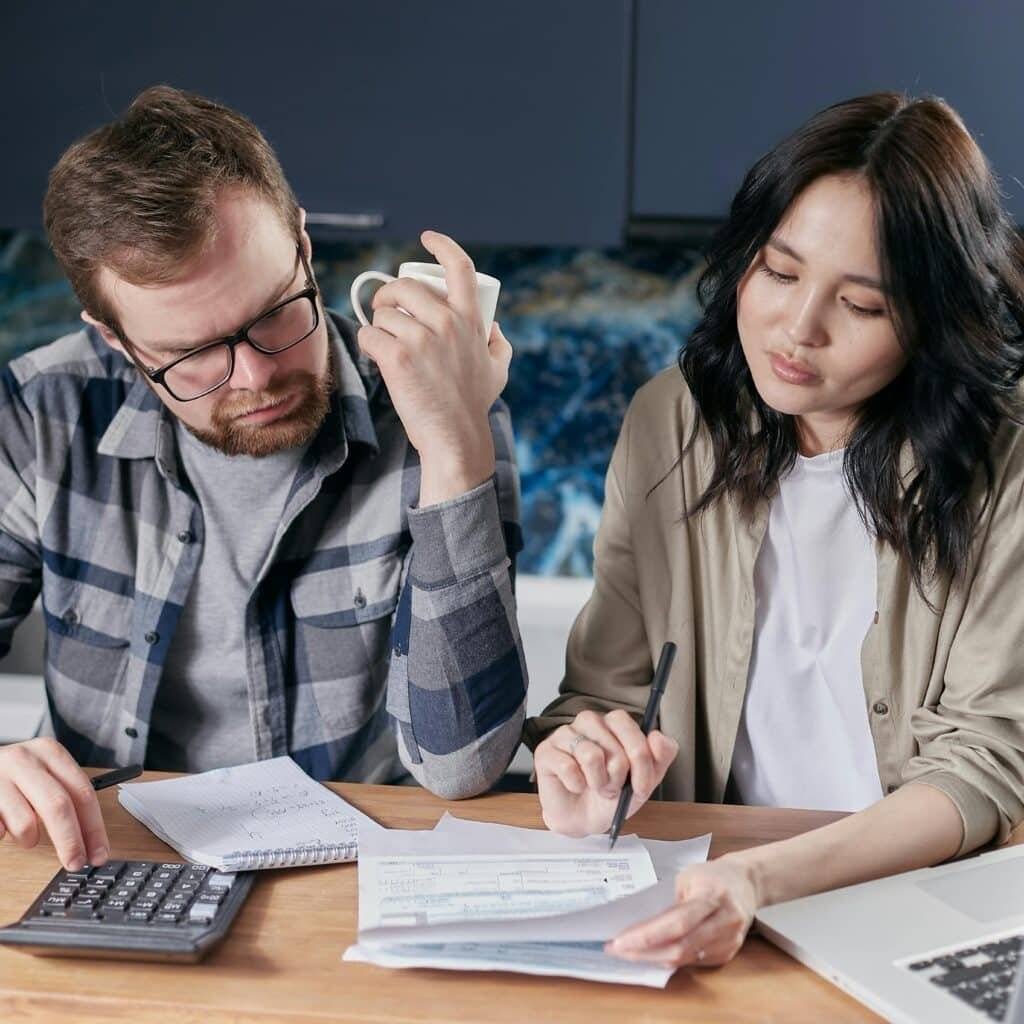 A couple reviewing household bills and budget using a calculator and laptop at their kitchen table.